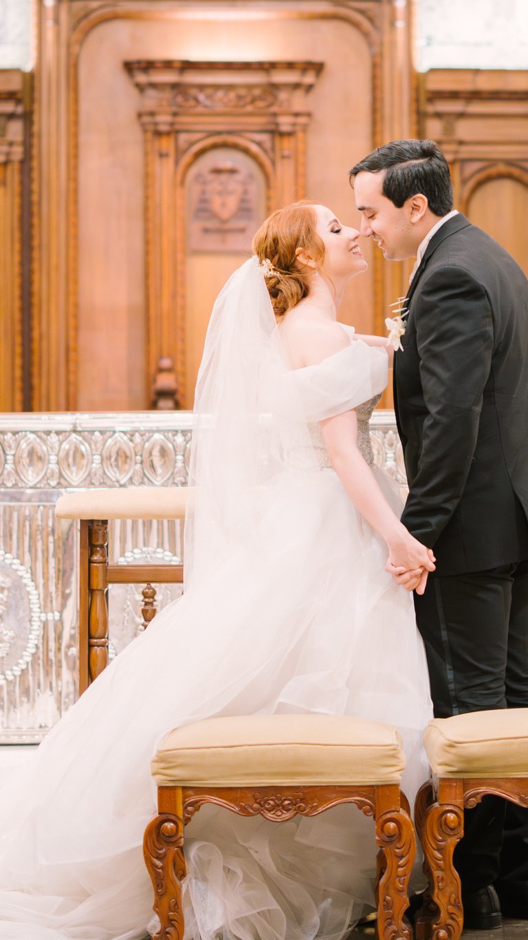 Novios tomados de la mano dándose un beso en el altar durante la ceremonia religiosa, con la novia en vestido blanco y el novio en traje negro.