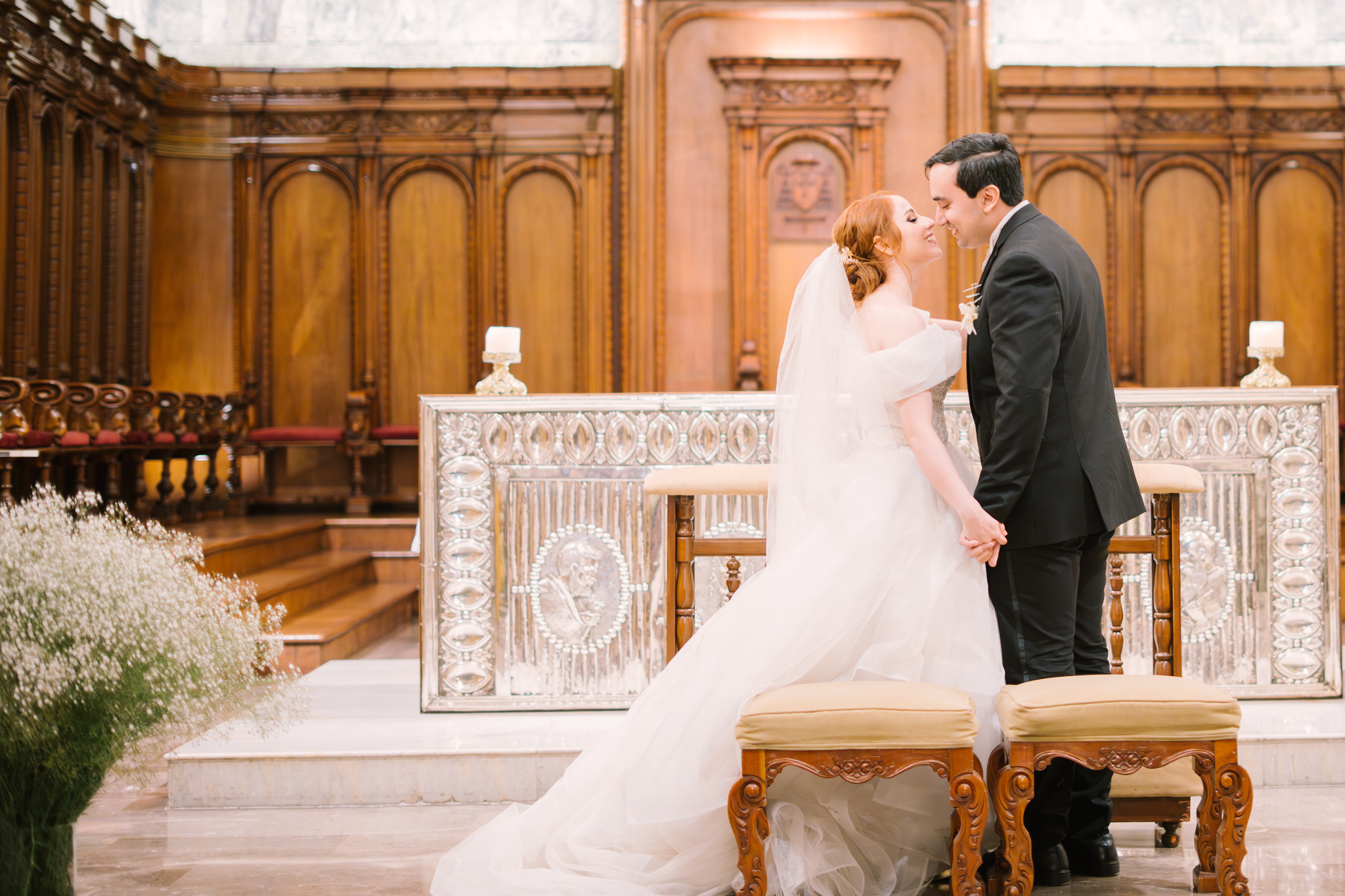 Novios tomados de la mano dándose un beso en el altar durante la ceremonia religiosa, con la novia en vestido blanco y el novio en traje negro.