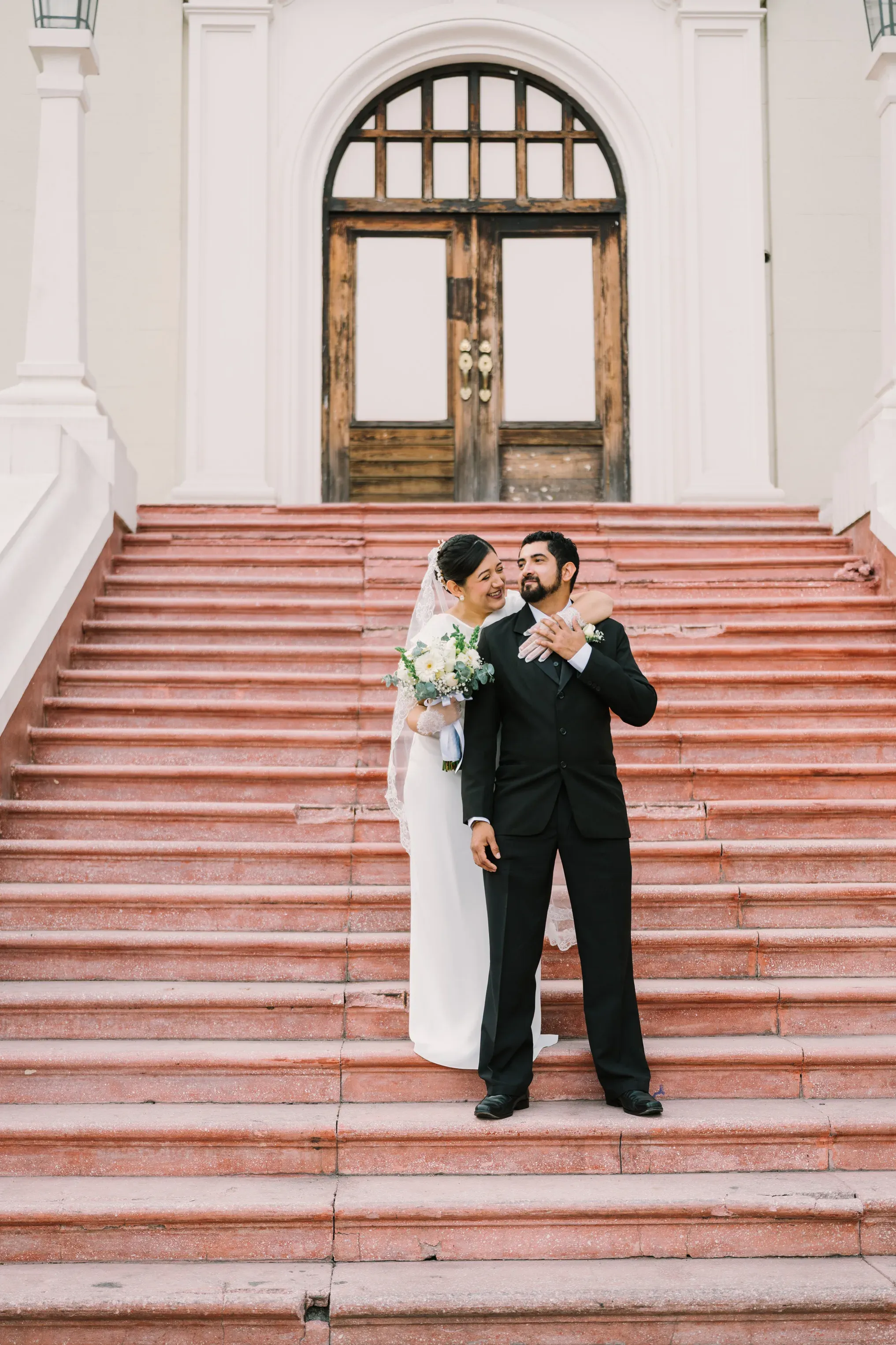 Amy y Jorge abrazados en las escaleras rojas del LABNL frente al edificio histórico blanco del centro de Monterrey