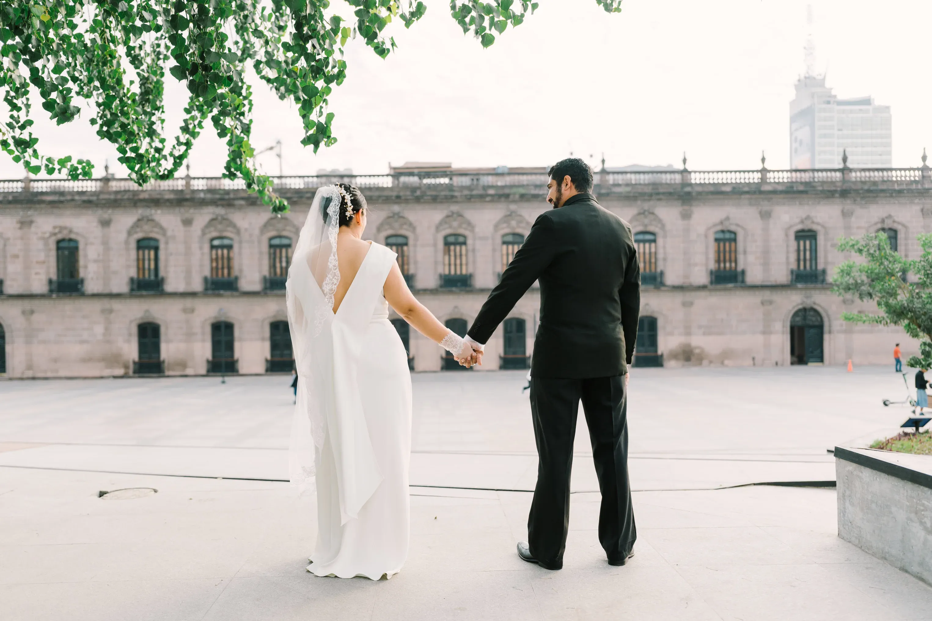 Vista trasera de Amy y Jorge tomados de la mano frente a la majestuosa fachada del Palacio de Gobierno de Nuevo León, en la Macroplaza del centro de Monterrey. Ramas con hojas verdes enmarcan la escena en la parte superior, mientras un edificio moderno asoma al fondo.
